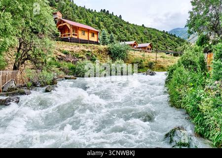 Rustikale Holzhütten auf einem Hügel überblicken einen schnell fließenden Bergfluss, umgeben von üppigen grünen Bäumen und Vegetation. Mountain River in Alamedin Go Stockfoto