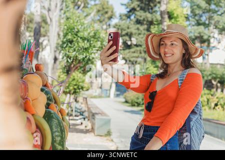 Lächelnde Frau mit Sommerhut macht ein Selfie mit ihrem Smartphone, während sie einen lebhaften Markt im Freien erkunden, umgeben von frischen Produkten und grünem du Stockfoto