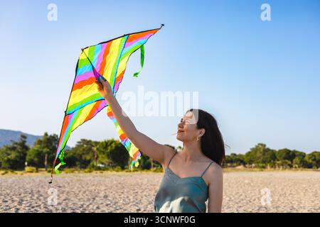 Eine fröhliche Mutter genießt einen sonnigen Tag am Strand und fliegt einen lebendigen, farbenfrohen Drachen vor einem klaren blauen Himmel. Ihr Lächeln spiegelt die unbeschwerte Freude des wider Stockfoto