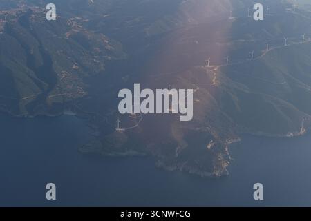 Atemberaubender Blick aus der Vogelperspektive auf Griechenlands Küstenlandschaft mit üppigen grünen Bergen und Windturbinen auf Graten, unter einem ruhigen blauen Himmel aus der Luft Stockfoto
