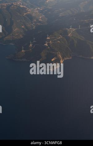 Blick aus der Vogelperspektive auf Griechenlands Küstenlandschaft mit zerklüfteten Klippen, üppigem Grün und mehreren Windturbinen im Gelände, alles unter dem weichen l Stockfoto