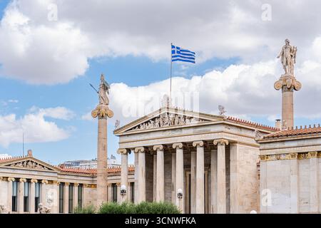 Neoklassizistische Architektur der Athener Akademie mit griechischer Flagge, die unter blauem Himmel winkt. Die ikonischen Säulen und Statuen veranschaulichen die historische Größe Stockfoto
