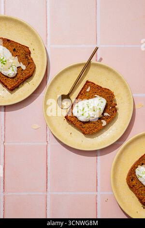 Draufsicht von drei Karottenkuchenscheiben mit Creme-Belag auf gelben Tellern, vor einem rosa gefliesten Hintergrund. Ein Löffel liegt neben einer Schicht, hochlig Stockfoto