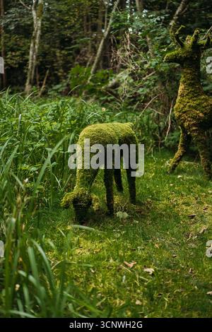 Zwei bezaubernde Hirschskulpturen, die sorgfältig mit üppigem Moos bedeckt sind, stehen inmitten von grünem Laub in einer ruhigen Waldlandschaft und harmonieren harmonisch Stockfoto