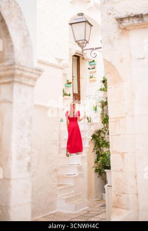 Eine Frau in rotem Kleid bestieg Steintreppen in der malerischen Stadt Cisternino, Apulien, Italien, umgeben von traditionellen weißen Gebäuden und üppigem Grün Stockfoto