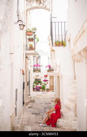 Eine Frau in rotem Kleid sitzt friedlich in einer malerischen Gasse von Cisternino, Apulien, umgeben von weiß getünchten Wänden und lebendigen Blumen, die das Land fängt Stockfoto