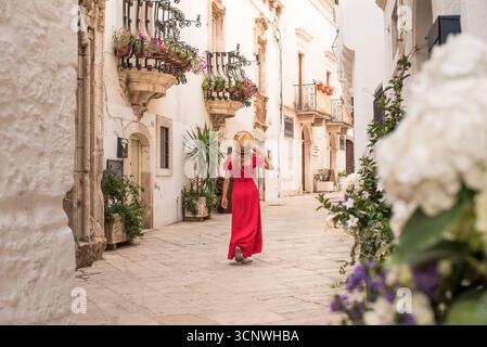 Eine Frau in rotem Kleid schlendert durch eine charmante, enge Straße in Locorotondo, Apulien, Italien, umgeben von historischen Gebäuden, die mit bunten Farben geschmückt sind Stockfoto