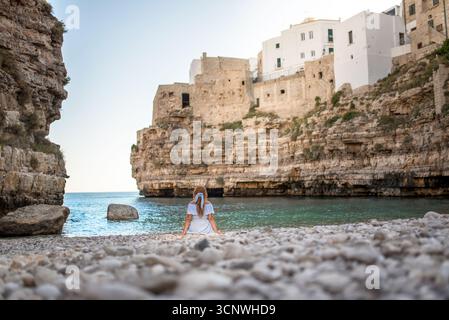 Eine friedliche Szene in Polignano a Mare, Apulien, Italien, mit einer Frau, die an einem Kiesstrand mit Blick auf das türkisfarbene Meer sitzt, umgeben von der Stadt Stockfoto