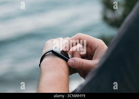 Nahaufnahme einer Hand eines Mannes, die mit einer Smartwatch vor einer ruhigen Kulisse des Meeres interagiert. Das Bild spiegelt eine Mischung aus Technologie, Natur und Passform wider Stockfoto