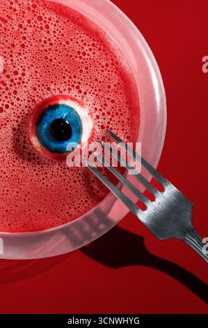 Ein gruseliges Halloween-Set mit einer realistischen Augapfelstütze, die in einer roten Flüssigkeit schwimmt, die Blut ähnelt, und einer Metallgabel, die darüber steht, wird mit Akzent gesetzt Stockfoto