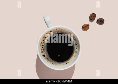 Ein Blick von oben auf eine weiße Tasse gefüllt mit schwarzem Kaffee, begleitet von verstreuten Kaffeebohnen auf einem hellbeigen Hintergrund, was ein minimalistisches und Stockfoto
