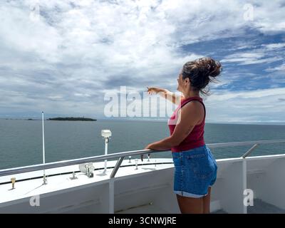 Eine Frau steht auf einem Boot in Cairns, Australien, und zeigt auf eine Insel am Horizont unter einem bewölkten Himmel, die Abenteuer und Entdeckungen auf dem Meer verkörpert Stockfoto