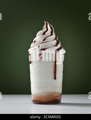 Ein verführerischer Schuss cremiges Vanilleeis mit reichhaltigem Schokoladenbesatz in einer transparenten Tasse vor schlichtem grünem Hintergrund, das einen Ref bietet Stockfoto