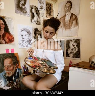 Eine junge Künstlerin taucht in ihr persönliches Atelier in die Malerei ein. Umgeben von ihrem Kunstwerk nutzt sie geschickt eine Palette, um ihr kreativ zu wirken Stockfoto