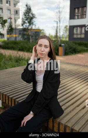 Die junge Biologin mit schwarzer Jacke und weißem Oberteil sitzt auf einer Holzbank, umgeben von urbanem Grün, die eine Mischung aus Natur und urb widerspiegelt Stockfoto
