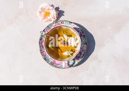 Eine wunderschön gefertigte Tasse Kräutertee sitzt elegant auf einer Untertasse mit Blumenmuster, begleitet von einer zarten rosa Blume. Die Komposition strahlt warm aus Stockfoto