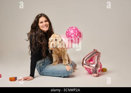Eine junge Frau lächelt fröhlich, während sie einen Geburtstag mit ihrem Cocker Spaniel feiert. Die dekorierte Einrichtung umfasst pinke Ballons und ein großes Metallic-Design Stockfoto