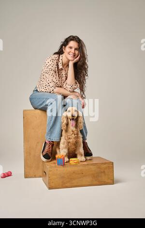 Eine Frau sitzt auf einer Holzkiste neben einem glücklichen Cocker Spaniel. Sie trägt lässige Jeans und ein Polka Dot-Shirt, was eine entspannte und freundliche Atmosphäre schafft. Stockfoto