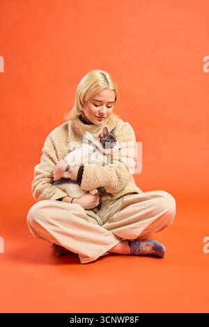 Eine Frau mit blonden Haaren sitzt im Kreuz in einem Studio, trägt einen gemütlichen Pullover und eine entspannte Hose. Sie hält liebevoll eine niedliche Katze in ihren Armen Agi Stockfoto