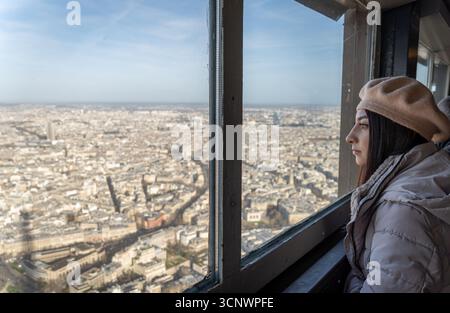 Eine Frau in beigefarbenem Baskenmütze und Wintermantel blickt vom Eiffelturm aus über die Skyline von Paris an einem ruhigen Morgen, eingetaucht in die atemberaubende Stockfoto
