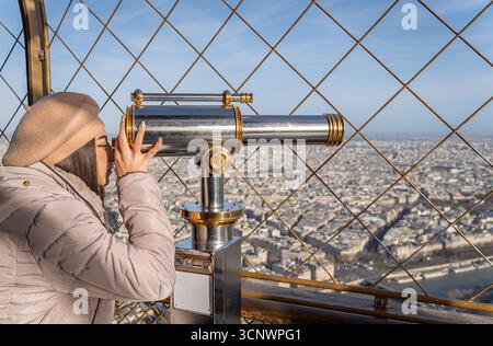 Eine Frau in beigefarbenem Baskenmütze und Mantel blickt durch ein Teleskop vom Eiffelturm aus und nimmt den Blick auf Paris am Morgen auf Stockfoto