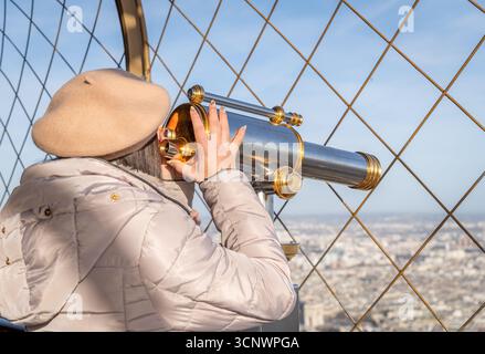 Eine Frau in beigefarbenem Baskenmütze und Wintermantel schaut durch ein Teleskop auf den Eiffelturm und genießt einen ruhigen Blick auf Paris am Morgen vom berühmten Landma aus Stockfoto