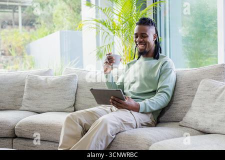 Afroamerikaner, der auf einem grauen Sofa im Wohnzimmer sitzt und einen Keramikbecher hält und ein Tablet benutzt Stockfoto
