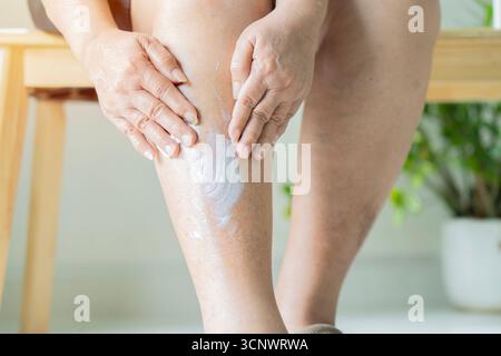 Zugeschnittene Ansicht einer älteren Frau, die Lotion auf ihr Bein aufträgt, während sie zu Hause auf einem Holzstuhl sitzt. Senior Healthy Konzept. Stockfoto