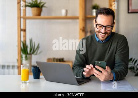 Selbstbewusster, lächelnder Geschäftsmann mit Smartphone im Heimbüro. Überprüfen neuer E-Mails, Videoanrufe Stockfoto