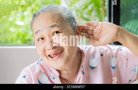 Glückliche ältere asiatische Frau sitzt neben dem Fenster und hält ihre Hand nahe dem Ohr mit Hörproblemen. Senior Healthy Konzept. Stockfoto