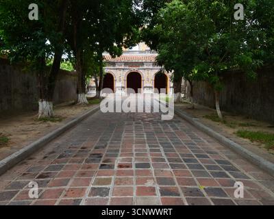 Steinweg, der zu einem königlichen Tempel in Hue führt - Ein flacher Ausschnitt eines traditionellen, gekachelten Weges, flankiert von Bäumen und Mauern, führt zu einem mehrbogigen G Stockfoto