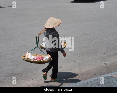 Traditioneller Straßenverkäufer mit Schulterstange - Ein Straßenverkäufer mit einem traditionellen konischen Hut trägt Waren in Körben, die auf einer Schulterstange balanciert sind. Stockfoto