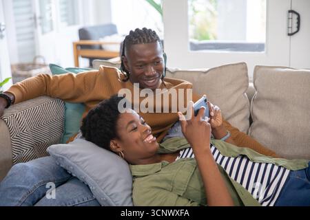 afroamerikanisches Paar, das sich auf dem Sofa mit Kissen im Wohnzimmer entspannt und den Bildschirm des Smartphones ansieht Stockfoto