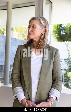 Eine ältere Frau, die neben der Schiebetür auf der Terrasse steht, in einem Oliven-Blazer und einem T-Shirt, und gard anstarrt Stockfoto