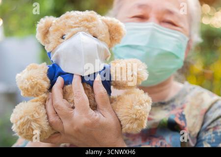 Glückliche ältere asiatische Frau, die einen kleinen braunen Teddybären hält, der auch eine medizinische Maske trägt. Senior Healthy Konzept. Selektiver Fokus. Stockfoto