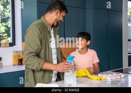 Indischer Vater mit Sohn, der auf der Arbeitsplatte in der Küche steht und Reiniger sprüht, während er Handschuhe trägt Stockfoto