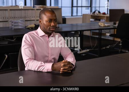 afroamerikanischer Mann sitzt am Schreibtisch und schließt Hände in einem modernen Büro mit Telefon Stockfoto