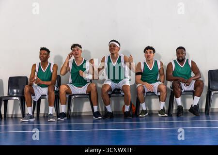 Verschiedene männliche Teamkollegen jubeln auf Stühlen auf dem Basketballplatz in Trikots mit Basketball Stockfoto