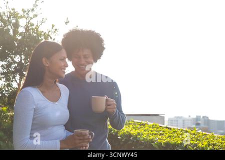 Verschiedene Paare halten Kaffeetassen und lächeln einander auf der Dachterrasse mit der Skyline der Stadt an Stockfoto