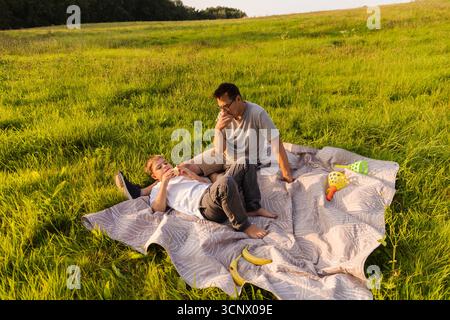 Ein Vater und sein kleiner Sohn entspannen sich auf einer weichen Decke in einem üppigen, grünen Feld. Sie teilen einen freudigen Moment voller Lachen und Snacks auf einer warmen, sonnigen Atmosphäre Stockfoto