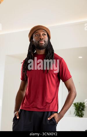 Afroamerikaner steht im Flur mit rotem Hemd, schwarzer Hose, brauner Beanie in der Nähe von Topfpflanzen Stockfoto