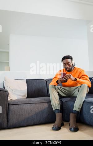 Afroamerikanischer Mann, der auf Sofa im Wohnzimmer sitzt und ein weißes Headset trägt, das kastanienbraunes Smartphone hält Stockfoto