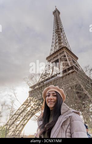 Von unten aus lächelt eine Frau in einem beigefarbenen Baskenmütze und Mantel vor dem berühmten Eiffelturm an einem frischen Wintermorgen in Paris, Capturin Stockfoto