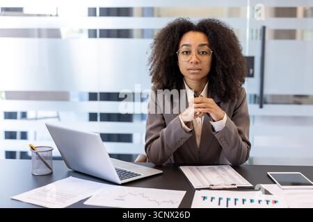 Porträt einer ernsten jungen afroamerikanischen Frau, die am Schreibtisch im Büro sitzt, mit Notizbuch und Dokumenten und selbstbewusst in die Kamera blickt. Stockfoto