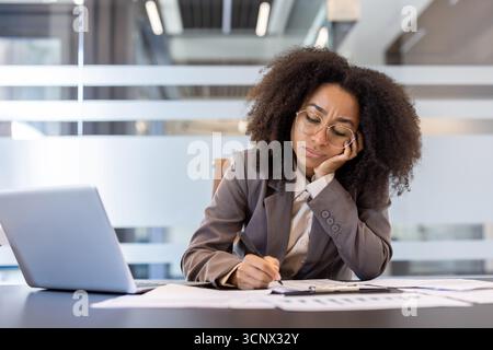 Frustrierte und müde afroamerikanische Frau, die im Anzug am Schreibtisch sitzt. Sie legte ihren Kopf auf die Hand und langweilte sich von ihrer Arbeit. Stockfoto