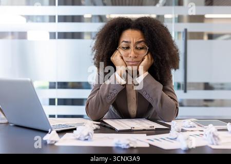 Müde junge afroamerikanische Frau, die am Schreibtisch im Büro sitzt und frustriert und verärgert über zerknittertes Papier, gescheiterte Arbeit und Ideen aussieht. Stockfoto