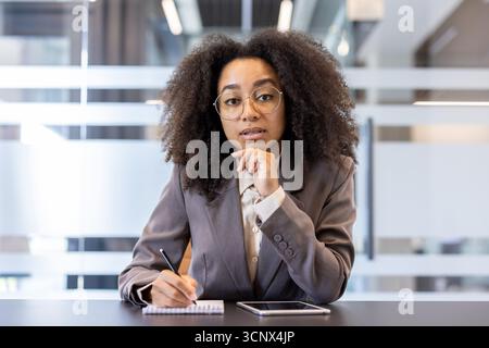 Porträt einer jungen afroamerikanischen Geschäftsfrau in Anzug und Brille, die am Schreibtisch im Büro vor der Kamera sitzt, einen Stift hält und Notizen macht. Stockfoto