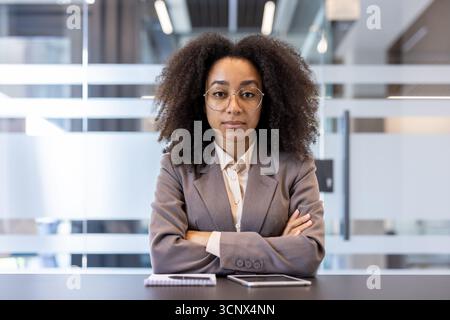 Porträt einer jungen seriösen afroamerikanischen Geschäftsfrau in Anzug und Brille, die ihre Arme auf der Brust kreuzt, am Schreibtisch im Büro sitzt und selbstbewusst in die Kamera schaut. Stockfoto