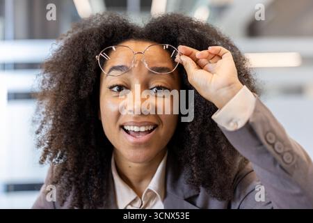 Nahporträt einer afroamerikanischen Frau mit lockigem Haar und Anzug in einem Büro, die mürrisch in die Kamera blickt und eine Brille auf ihrem Gesicht hält. Stockfoto