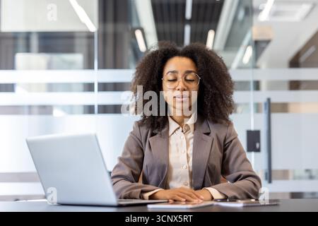 Junge afroamerikanische Geschäftsfrau im Anzug, die am Schreibtisch im Büro sitzt, die Augen geschlossen hat und tief atmet. Stockfoto